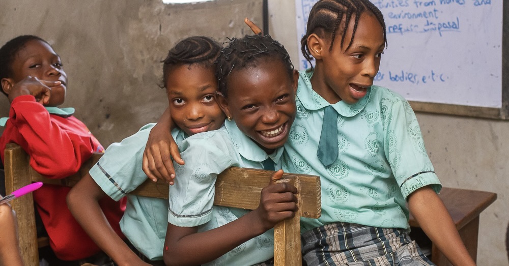 Image of school-aged girls hugging and smiling in a classroom. 